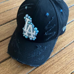 Peut inclure: Casquette de baseball noire avec des détails usés. La casquette présente les lettres "LA" dans un design stylisé, agrémentées d'accents bleu clair et argenté. Des ornements supplémentaires en forme d'étoiles bleues sont dispersés sur la casquette.