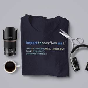 May include: A dark blue t-shirt with the code "import tensorflow as tf" printed on it. The code is written in a light blue, green, and yellow gradient. The t-shirt is folded and laid flat on a grey surface.