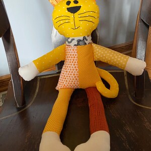 May include: A handmade stuffed cat doll with a yellow head, ears, and arms. The cat has a black stitched face, a patterned collar, and a patchwork body in orange and yellow. The legs are yellow and brown, with white feet.