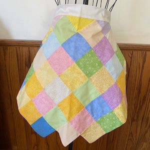 May include: A patchwork apron with a white waistband. The apron is made of squares of different pastel colors, including yellow, pink, blue, and green.