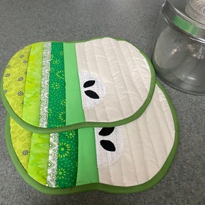 May include: Two green and white quilted pot holders shaped like apples. The pot holders have a green border and a white center with a black apple seed design.