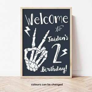 Puede incluir: Un cartel imprimible en blanco y negro con una mano de esqueleto haciendo un signo de paz. El cartel dice "Bienvenido a Jaiden's 2nd Birthday!" con rayos en ambos lados del n&uacute;mero 2.