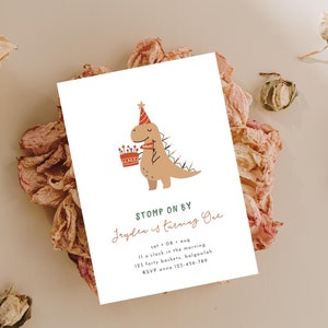 May include: A white card with a cartoon dinosaur wearing a party hat and holding a birthday cake. The text on the card reads "STOMP ON BY Jayden is turning One" with details about the party.