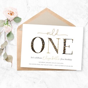 May include: A white invitation card with gold text and a brown envelope. The card reads "wild ONE" with the "O" in a zebra print and the "N" and "E" in a leopard print. The text below says "let's celebrate Elizabeth's first birthday".