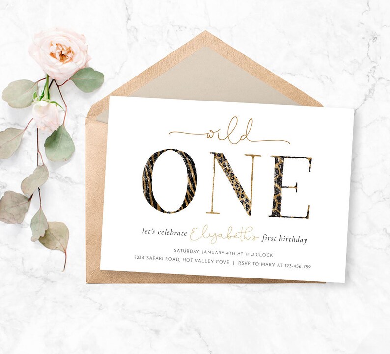 May include: A white invitation card with gold text and a brown envelope. The card reads "wild ONE" with the "O" in a zebra print and the "N" and "E" in a leopard print. The text below says "let's celebrate Elizabeth's first birthday".