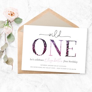 May include: A white invitation card with the words "wild ONE" in black script and animal print lettering. The card is for a first birthday and includes event details. A kraft paper envelope and pink flowers are in the background.