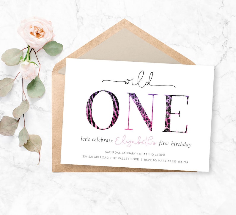 May include: A white invitation card with the words "wild ONE" in black script and animal print lettering. The card is for a first birthday and includes event details. A kraft paper envelope and pink flowers are in the background.
