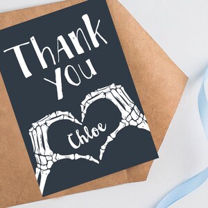 Puede incluir: Una tarjeta de agradecimiento azul oscuro con manos de esqueleto blancas formando un coraz&oacute;n con el nombre "Chloe" en su interior. El texto "Gracias" est&aacute; escrito en blanco en la tarjeta.