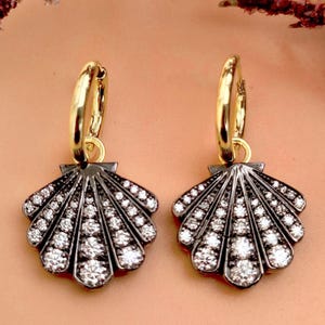 Może przedstawiać: Para złotych kolczyków koła z motywem muszli. Zawieszki w kształcie muszli są ciemnoszare z białymi kamieniami szlachetnymi. Kolczyki są prezentowane na brzoskwiniowym tle z bordowymi liśćmi.