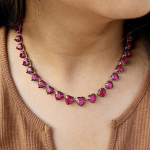 May include: A necklace featuring heart-shaped, deep pink gemstones. The necklace is worn against a brown top. The gemstones are closely set together, creating a continuous line of color around the neck. The necklace is a piece of jewelry.