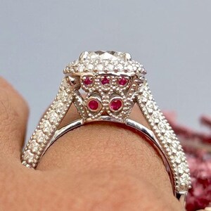 May include: A silver engagement ring with a large center stone surrounded by smaller diamonds. The ring has a halo of red gemstones around the center stone.
