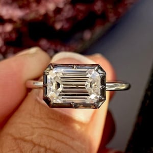 Puede incluir: Un anillo de plata con un diamante de talla esmeralda. El anillo está engastado en una banda de oro blanco y tiene un halo negro alrededor del diamante. El anillo está en una caja de terciopelo verde con el texto "Kohinoor Boutique" en ella.
