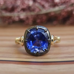 May include: A gold ring featuring a large, oval, deep blue gemstone. The gemstone is set in a dark metal bezel. The ring rests on a light brown wooden surface, with a blurred floral arrangement behind it.