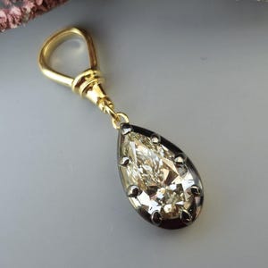 Op de afbeelding: Een goudkleurige hanger met een druppelvormige, heldere steen. De steen is gezet in een donkere metalen zetting. De hanger hangt aan een gouden lus. Roze bloemen zijn op de achtergrond.