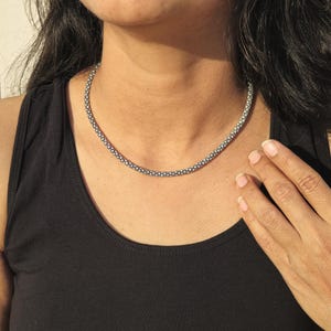 May include: A silver-colored beaded necklace is worn around the neck. The necklace is made of small, round beads. The person is wearing a black tank top. The person's hand is resting on their chest.