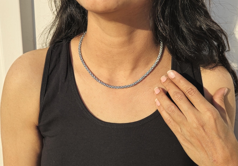 May include: A silver-colored beaded necklace is worn around the neck. The necklace is made of small, round beads. The person is wearing a black tank top. The person's hand is resting on their chest.