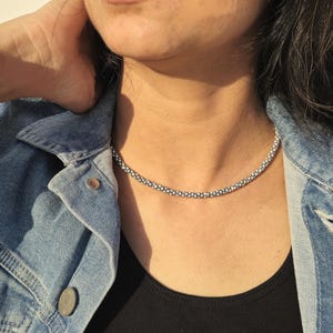 May include: A silver beaded necklace resting on a person's neck. The necklace is composed of small, round beads. The person is wearing a black top and a denim jacket. The jacket has silver buttons.