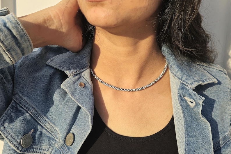 May include: A silver beaded necklace resting on a person's neck. The necklace is composed of small, round beads. The person is wearing a black top and a denim jacket. The jacket has silver buttons.