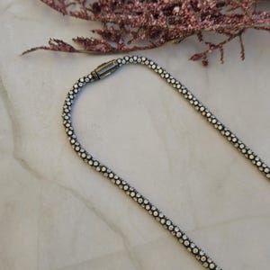 May include: A silver-toned necklace with a beaded design, featuring a magnetic clasp. The necklace is displayed on a marble surface, with a sprig of dried pink flowers in the background. The necklace is a jewelry item.