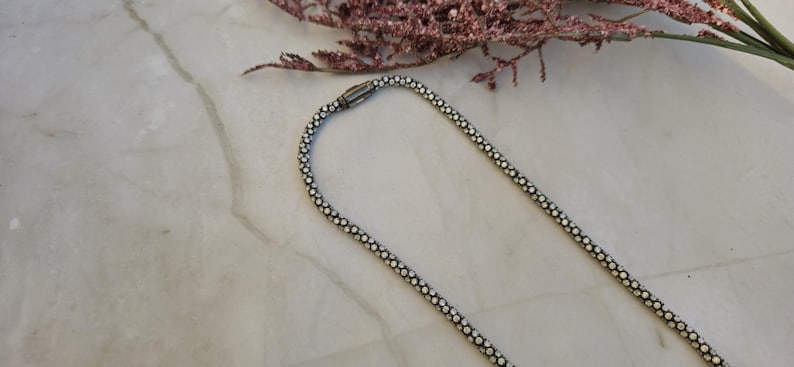 May include: A silver-toned necklace with a beaded design, featuring a magnetic clasp. The necklace is displayed on a marble surface, with a sprig of dried pink flowers in the background. The necklace is a jewelry item.