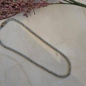 May include: A silver-toned necklace with a delicate beaded design, lying on a marble surface. The necklace is composed of small, round beads linked together to form a chain. A sprig of dried flowers is in the background.
