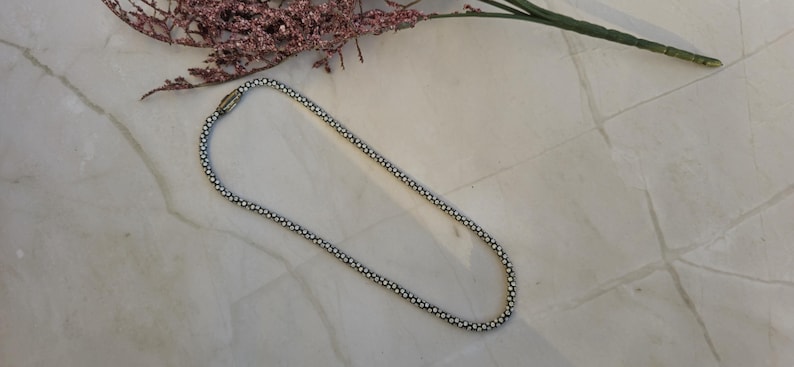 May include: A silver-toned necklace with a delicate beaded design, lying on a marble surface. The necklace is composed of small, round beads linked together to form a chain. A sprig of dried flowers is in the background.