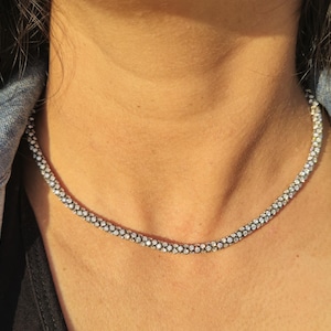 May include: A silver-colored beaded necklace. The necklace is worn over a black top and a denim jacket. The necklace is made of small, round beads that are closely spaced together, creating a continuous, textured appearance.