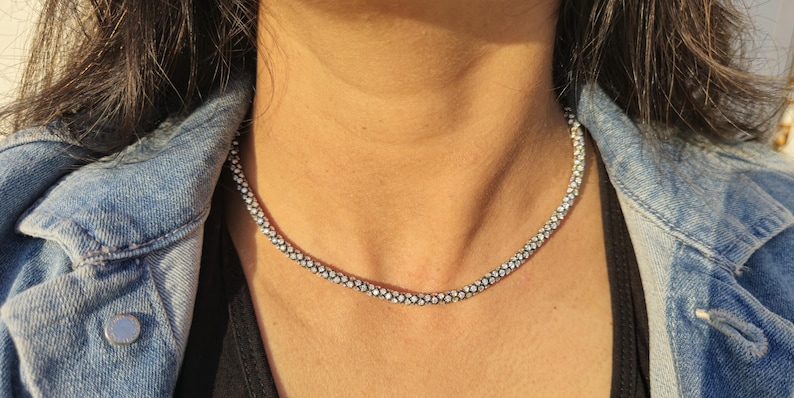 May include: A silver-colored beaded necklace. The necklace is worn over a black top and a denim jacket. The necklace is made of small, round beads that are closely spaced together, creating a continuous, textured appearance.