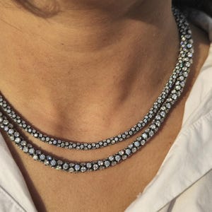 May include: Two-strand necklace with a dark metal setting and numerous small, sparkling stones. The necklace is worn over a white collared shirt. The stones appear to be clear and round, creating a dazzling effect.