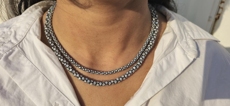 May include: Two-strand necklace with a dark metal setting and numerous small, sparkling stones. The necklace is worn over a white collared shirt. The stones appear to be clear and round, creating a dazzling effect.