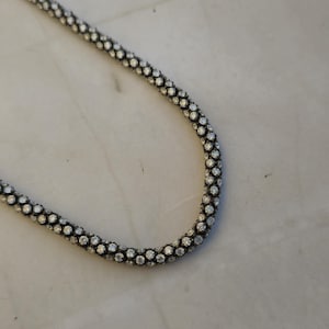 May include: A silver-colored necklace with a beaded design. The necklace is made of small, round beads that are closely spaced together, creating a textured surface. The necklace is displayed on a white surface.