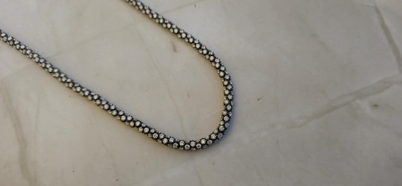 May include: A silver-colored necklace with a beaded design. The necklace is made of small, round beads that are closely spaced together, creating a textured surface. The necklace is displayed on a white surface.