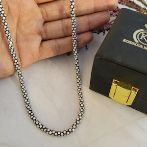 May include: A silver-toned necklace with a beaded design is displayed next to a black jewelry box with a gold clasp. The box has the text "KOHINOOR BOUTIQUE" and a logo. The necklace is draped over a hand.