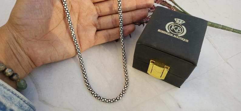 May include: A silver-toned necklace with a beaded design is displayed next to a black jewelry box with a gold clasp. The box has the text "KOHINOOR BOUTIQUE" and a logo. The necklace is draped over a hand.