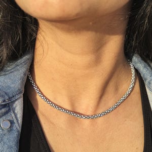 May include: A silver-colored necklace with a delicate, beaded design. The necklace is worn against a black top and a denim jacket. The necklace appears to be made of small, round beads.