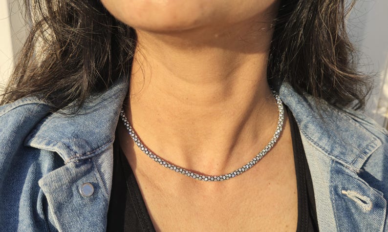 May include: A silver-colored necklace with a delicate, beaded design. The necklace is worn against a black top and a denim jacket. The necklace appears to be made of small, round beads.