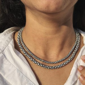 May include: A two-strand necklace with a silver-tone metal setting and numerous small, clear gemstones. The necklace is worn over a white collared shirt. The gemstones are closely set, creating a sparkling effect.