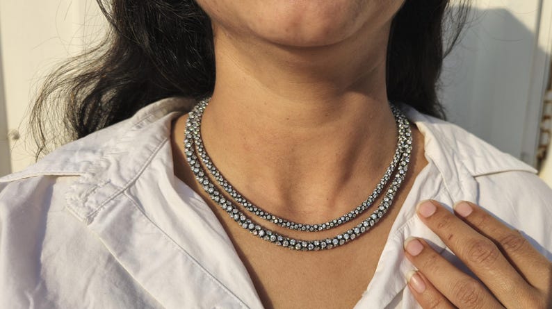 May include: A two-strand necklace with a silver-tone metal setting and numerous small, clear gemstones. The necklace is worn over a white collared shirt. The gemstones are closely set, creating a sparkling effect.