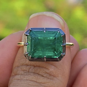 Peut inclure: Une bague ornée d'une grande pierre précieuse verte de taille émeraude rectangulaire, sertie dans une monture en métal foncé oxydé avec des griffes. L'anneau est composé d'un métal doré fin et poli, contrastant avec le sertissage sombre.