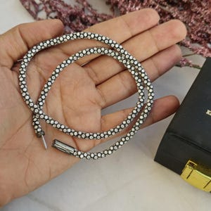 May include: A silver-toned necklace with small, clear gemstones is displayed. The necklace is coiled in a hand, next to a black box with gold hardware and the text "KOHINOOR BOUTIQUE".