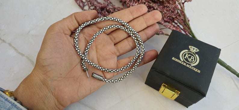May include: A silver-toned necklace with small, clear gemstones is displayed. The necklace is coiled in a hand, next to a black box with gold hardware and the text "KOHINOOR BOUTIQUE".