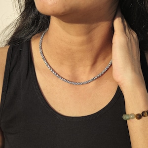 May include: A silver-toned beaded necklace and a beaded bracelet with brown and green stones. The necklace is a choker style, and the bracelet is worn on the wrist. The person is wearing a black tank top.