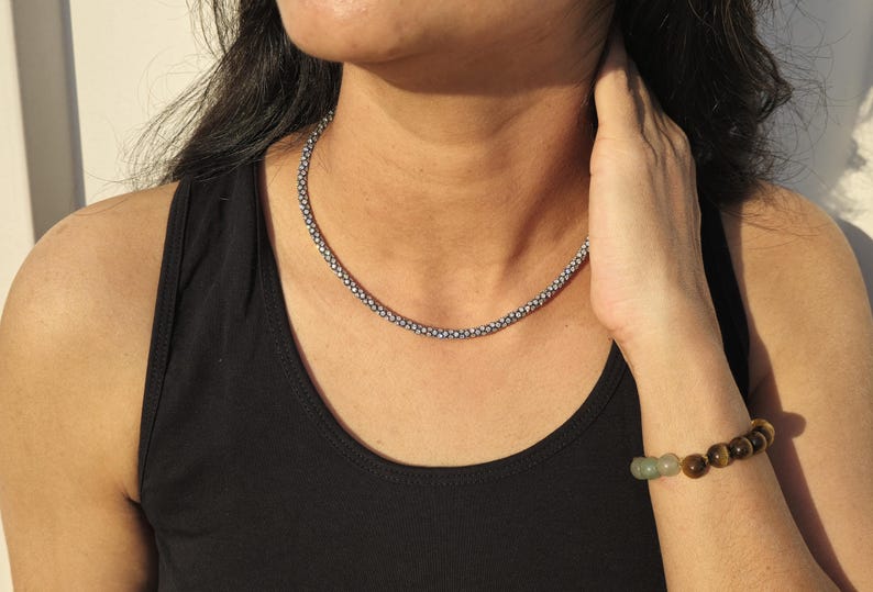 May include: A silver-toned beaded necklace and a beaded bracelet with brown and green stones. The necklace is a choker style, and the bracelet is worn on the wrist. The person is wearing a black tank top.