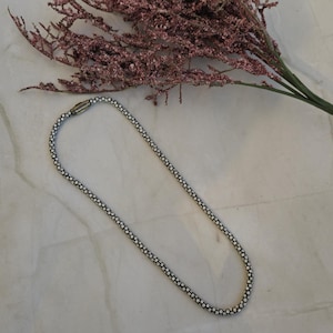 May include: A silver-toned necklace with a beaded design lies on a marble surface, next to a sprig of dried pink flowers. The necklace is a simple, elegant piece of jewelry.