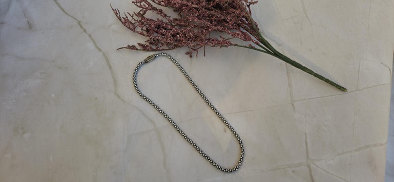 May include: A silver-toned necklace with a beaded design lies on a marble surface, next to a sprig of dried pink flowers. The necklace is a simple, elegant piece of jewelry.