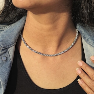 May include: A silver-colored beaded necklace is worn over a black top. The necklace is a choker style. The person is wearing a denim jacket. The image is taken in natural light.