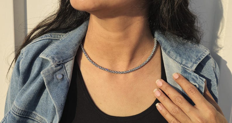 May include: A silver-colored beaded necklace is worn over a black top. The necklace is a choker style. The person is wearing a denim jacket. The image is taken in natural light.