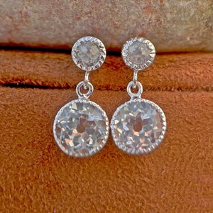 May include: A pair of silver dangle earrings with two clear round stones. The earrings are on a brown suede surface.