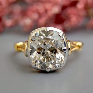 Op de afbeelding: Een grote, heldere, kussen geslepen diamanten ring gezet in een gouden en witgouden band. De ring heeft een delicaat, ingewikkeld ontwerp.