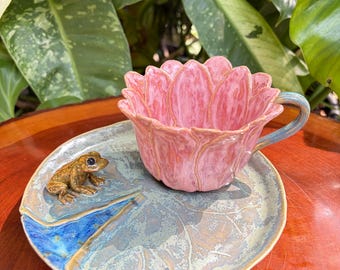 May include: A ceramic tea cup and saucer set featuring a pink lotus flower shaped cup with a blue handle. The saucer is designed as a green lily pad with a small, detailed brown frog figurine attached to the surface, finished in a glossy glaze.
